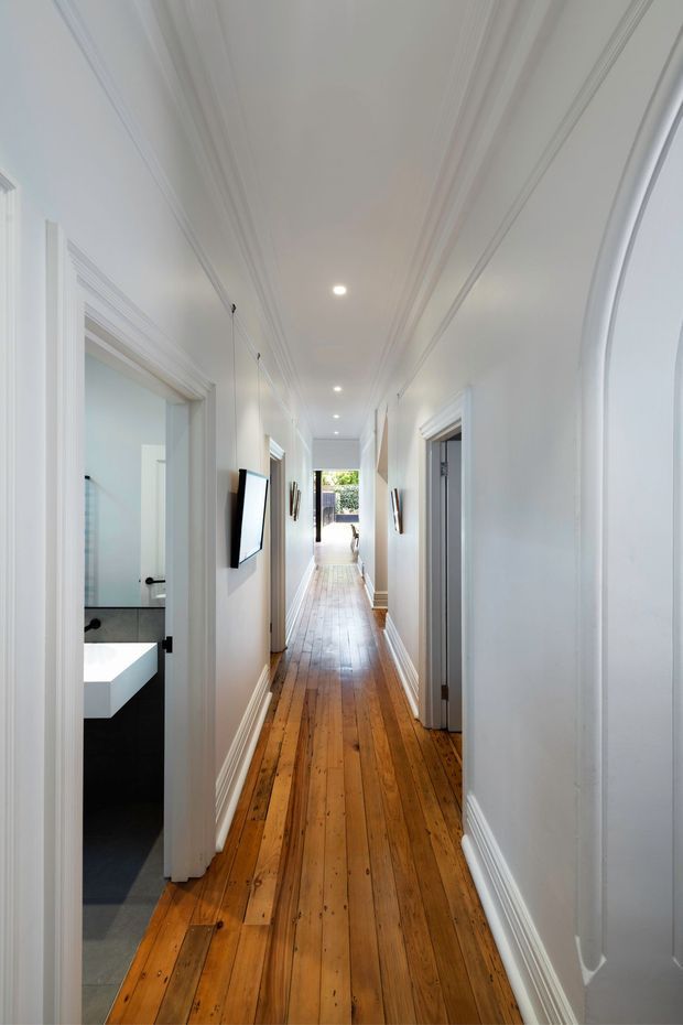 Bright, long residential hallway with polished warm timber floorboards, crisp white walls, elaborate decorative cornices and skirting, recessed ceiling downlights, and multiple white-trimmed doorways. A contemporary bathroom with black-and-white finishes is visible through the open doorway on the left, while the corridor leads toward a glazed opening at the far end.