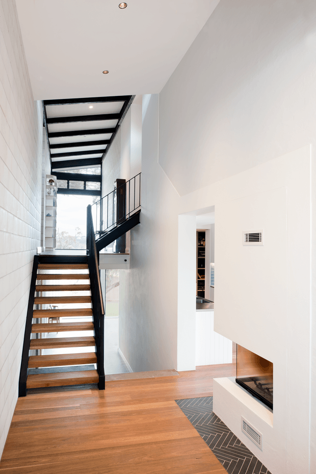A bright, contemporary stairway and landing with open timber stair treads, black metal stringers and balustrades, and a black-framed glazed roof or clerestory structure above. Warm timber flooring contrasts with white walls and a built-in white fireplace featuring a dark herringbone tile hearth. An opening reveals part of an adjacent room and fixed shelving.