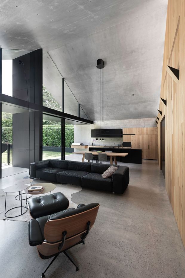 A spacious open-plan modern living and kitchen area with a double-height concrete ceiling, polished concrete floor, extensive black-framed glazing, and warm timber wall panelling. A black leather sectional sofa and lounge chair define the living zone, while a minimalist kitchen with a central dining island occupies the rear. The room opens visually to a leafy outdoor area and features slim black pendant lights and integrated cabinetry.
