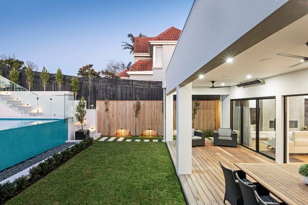 Contemporary outdoor entertaining area with a covered timber deck opening onto a landscaped courtyard and swimming pool. The space features white rendered walls, black-framed glazing, timber slat screening, recessed lighting, outdoor dining furniture, and a tiled pool with integrated steps. A neighbouring or existing house with a red tiled roof is visible beyond the courtyard.