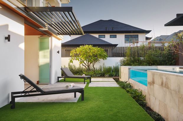 A contemporary landscaped courtyard beside a white rendered building, featuring a timber pergola with slatted shading, glass doors, lounge chairs, manicured lawn, planting, and a raised turquoise swimming pool enclosed by pale stone-clad walls. Neighbouring pitched-roof homes are visible beyond the courtyard.