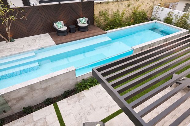Elevated view of a narrow rectangular swimming pool in a landscaped contemporary outdoor setting. The pool is surrounded by pale stone paving and concrete walls, with a dark timber chevron privacy screen and a small seating area at one end. A slatted metal pergola or balcony structure crosses the foreground, adding strong linear contrast.
