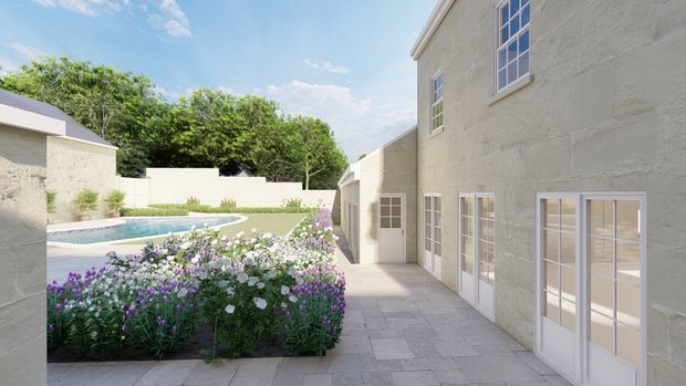 Sunlit courtyard beside a pale rendered masonry building, with multi-pane French doors and windows opening onto a paved walkway. A curved swimming pool and dense flowering garden border the courtyard, with mature trees beyond and a bright blue sky overhead.