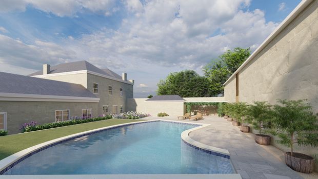 A landscaped courtyard-style outdoor pool area enclosed by pale rendered masonry buildings and walls. The curving swimming pool is bordered by light stone paving, raised planters, lawns and flowering garden beds, with a residential building and pitched roof visible beyond.