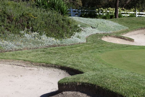 A landscaped golf course scene showing a closely mown putting green bordered by sculpted grass edges, pale sand bunkers, and dense ornamental planting. A white fence and mature vegetation appear in the background.