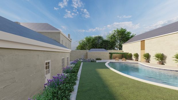A landscaped courtyard with a curved swimming pool surrounded by pale rendered masonry buildings with pitched dark roofs. The space includes manicured lawn, paved pool edging, planted garden beds, potted plants, and outdoor seating under a bright blue sky.