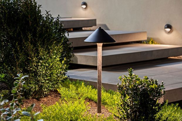 A landscaped outdoor entry or terrace area with layered concrete steps, integrated wall lighting, clipped shrubs, ornamental grasses, and a freestanding conical garden light. The design has clean geometric lines and a contemporary minimalist character.