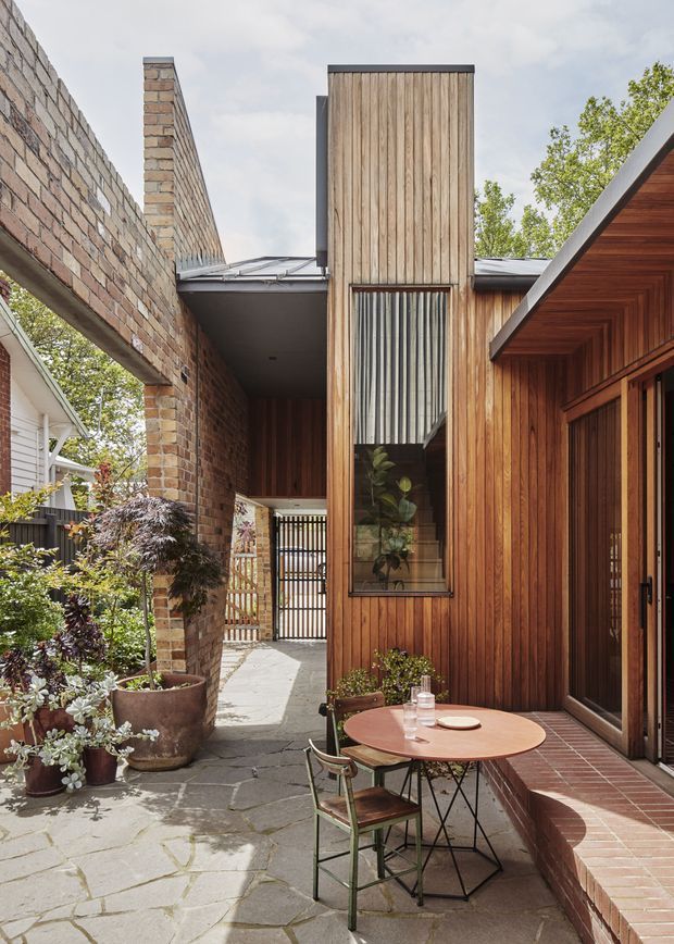 A narrow landscaped courtyard and outdoor dining terrace between brick and timber-clad residential volumes. The space features warm vertical timber wall cladding, exposed brick walls, a tall glazed opening with vertical screening, a pitched timber-lined eave, slate-like irregular stone paving, brick steps, and a small round café table with chairs. The composition has a contemporary courtyard character with strong material contrast and abundant potted greenery.