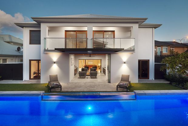 A contemporary two-storey white-rendered residence viewed from the rear, with a broad upper-level balcony, recessed ground-floor entertaining area, and large blue swimming pool in the foreground. Warm interior lighting, glass balustrades, dark accents, and neighbouring suburban homes are visible.