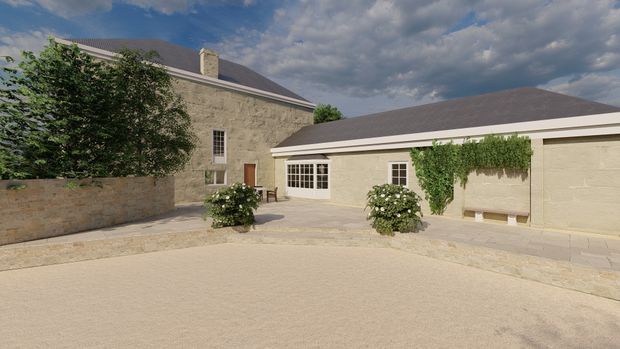 A stone-clad courtyard exterior formed by a taller gabled building and a lower adjoining wing. The buildings have muted grey pitched roofs, pale masonry walls, small multi-pane windows, a simple timber door, and climbing greenery. The broad paved courtyard includes low stone walls, clipped shrubs, and a light gravel or stone surface under a partly cloudy sky.