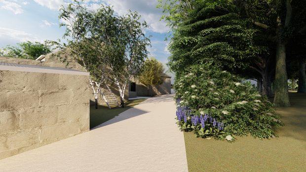 A landscaped residential walkway with a broad pale paved path winding between rendered masonry walls, clipped hedges, mature trees, and flowering garden beds. The scene has a quiet, refined garden character with contemporary low-rise structures partially visible in the background.