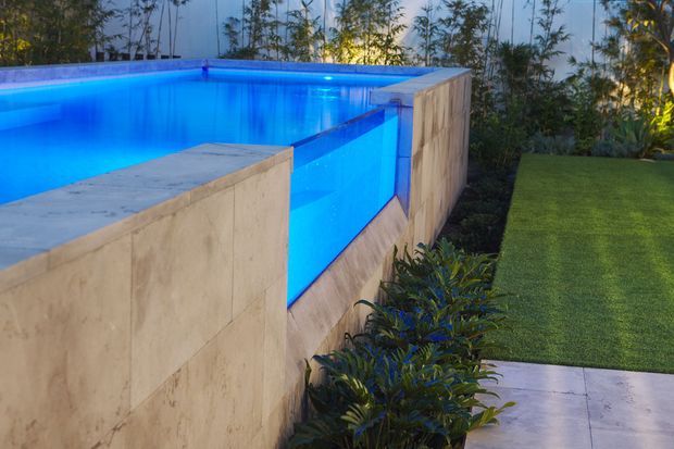 A contemporary outdoor swimming pool with vivid blue lighting, enclosed by low rendered or stone-clad walls. The pool edge and wall create a geometric composition beside a manicured lawn, tiled paving, and dense garden planting.