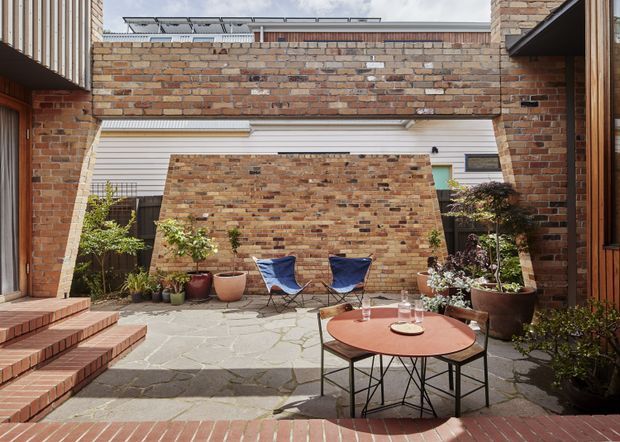 A brick-lined residential courtyard viewed from within the home, with stepped brick paving, irregular stone paving, raised brick walls, timber and brick facades, potted plants, two lounge chairs, and a round outdoor table. The enclosed space has a warm, textural modern courtyard character and includes a narrow strip of sky and roof detailing above.
