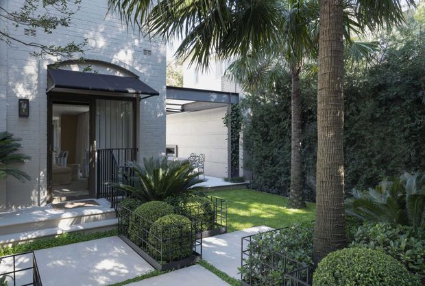 A landscaped residential entry courtyard viewed from outside, featuring a pale masonry facade, recessed glass entry door with a dark metal awning, concrete paths, black metal garden edging, clipped shrubs, palms, and dense screening greenery. A covered outdoor passage or carport-like structure is visible beyond the entry.