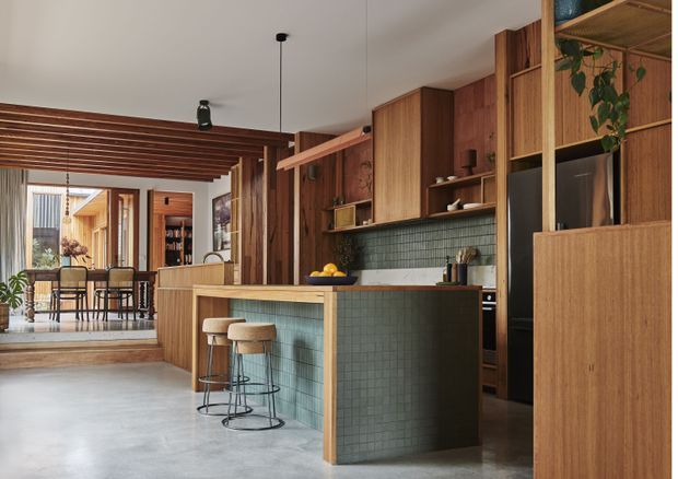 Warm, open-plan kitchen and dining area featuring extensive walnut-toned timber cabinetry and shelving, a green tiled kitchen island with timber benchtop, integrated appliances, and a concrete-look floor. A dining space is visible beyond the kitchen, separated by a timber slatted ceiling feature and a step-down transition. The space combines rich natural materials with an earthy, contemporary palette.