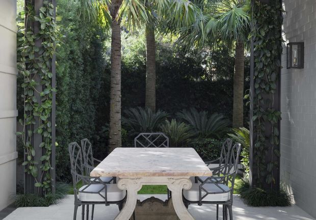 A refined outdoor dining terrace framed by climbing vines and lush tropical planting. A long pale stone dining table with ornate sculptural legs is surrounded by dark metal lattice-back chairs, set against a grey masonry wall and dense garden greenery.