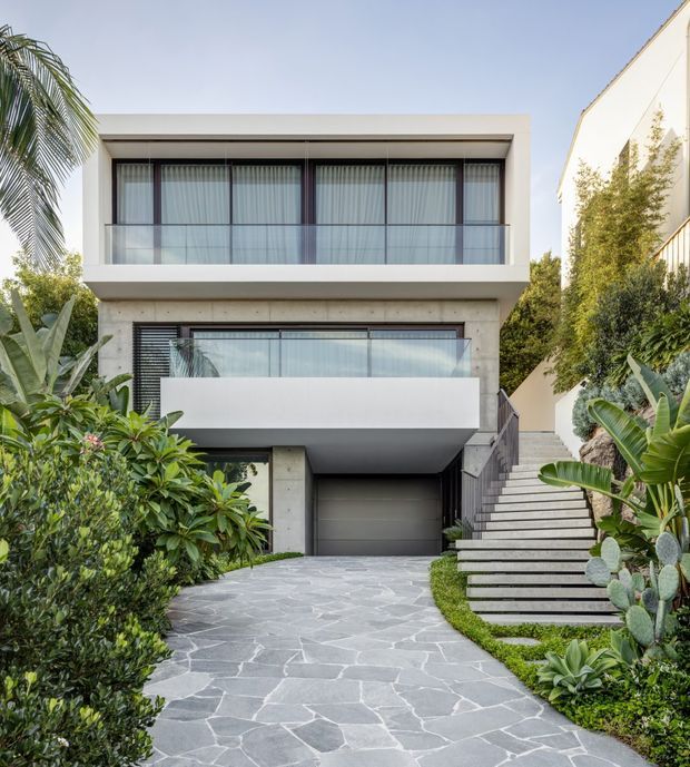 Front exterior view of a modern multi-storey residence with a minimalist rectangular form, broad glazed balconies, exposed concrete elements, dark window frames, and a concealed garage. A pale irregular stone-paved driveway leads through lush tropical landscaping, with a prominent stepped external stairway along the right side.