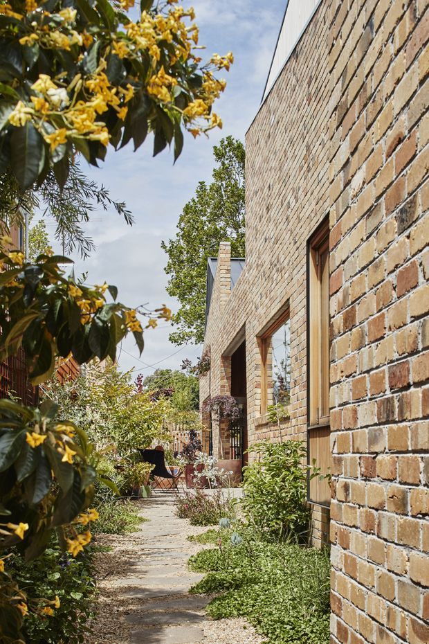 A narrow landscaped walkway runs alongside a long single-storey brick building. The facade features warm variegated brickwork, timber-framed windows and doors, and a simple pitched roofline. Dense flowering and evergreen planting borders the path, with a small outdoor seating area visible farther along. The image has a warm, natural residential architectural character.