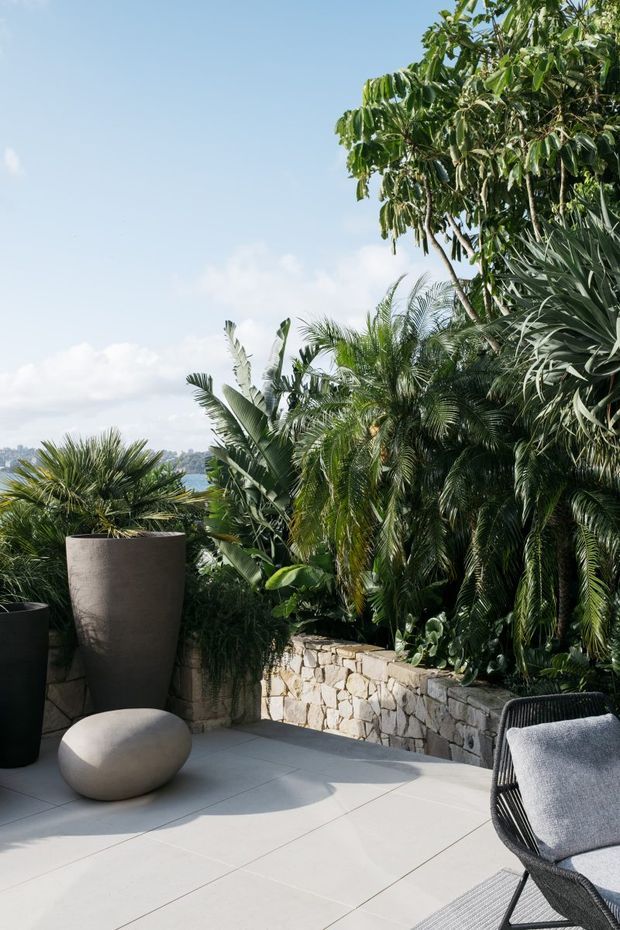 A sunlit outdoor terrace with pale large-format tiled paving, a low irregular natural-stone retaining wall, large dark planters, and a woven outdoor lounge chair. Dense tropical foliage forms a lush green backdrop, with distant urban scenery visible beyond the garden.