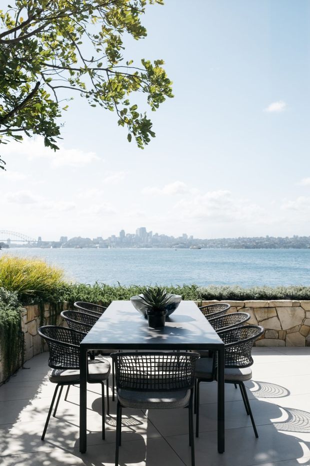 A bright waterfront outdoor dining terrace with a dark rectangular dining table and woven black chairs, overlooking a broad bay and distant city skyline. The terrace has pale tiled paving, a low irregular-stone retaining wall, clipped planting, and a large leafy tree providing partial shade. A landmark bridge is visible in the distant background.
