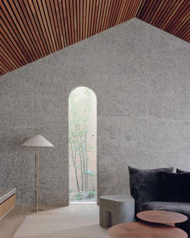 A refined contemporary living room with a high pitched ceiling lined in warm timber battens, textured grey concrete or plaster walls, and a tall arched window opening onto greenery. The room contains a dark upholstered sofa, sculptural round coffee tables, a simple floor lamp, and a small cylindrical side table. The palette is muted and earthy, with strong contrast between the dark furniture, pale floor, grey walls, and timber ceiling.