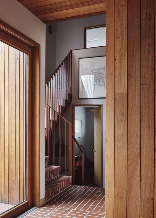 Interior view of a residential entry and stairway. A timber-framed glazed door sits beside a red brick-tiled floor and brick stair treads, with a dark red-painted balustrade leading upward. Warm timber ceiling boards and vertical timber wall cladding frame the space, with a framed artwork and a partially visible room beyond the stair landing.
