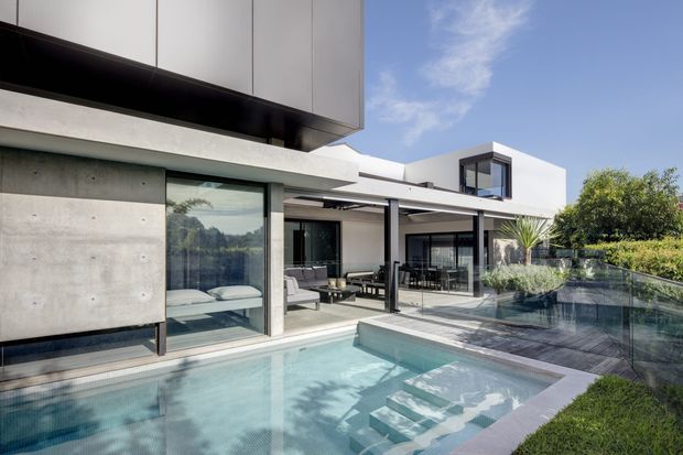 Contemporary multi-level home with crisp white rendered forms, exposed concrete walls, dark metal framing and extensive floor-to-ceiling glazing, opening onto a covered outdoor entertaining area and a compact swimming pool. The image is viewed from the landscaped pool area, with the home's terraces, glass balustrades and interior living spaces visible beyond.