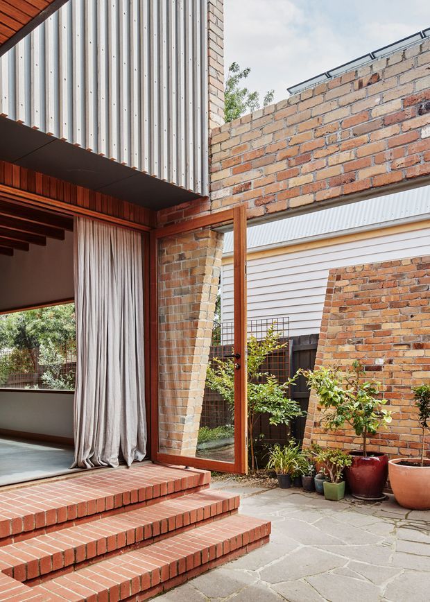 A transitional indoor-outdoor threshold with a large timber-framed glazed door opening onto a paved courtyard. The architecture features exposed red brick walls, corrugated metal cladding above, timber soffit detailing, broad brick steps, and irregular stone paving. A curtained interior room is visible on the left, while potted plants and a neighbouring weatherboard structure appear in the courtyard.