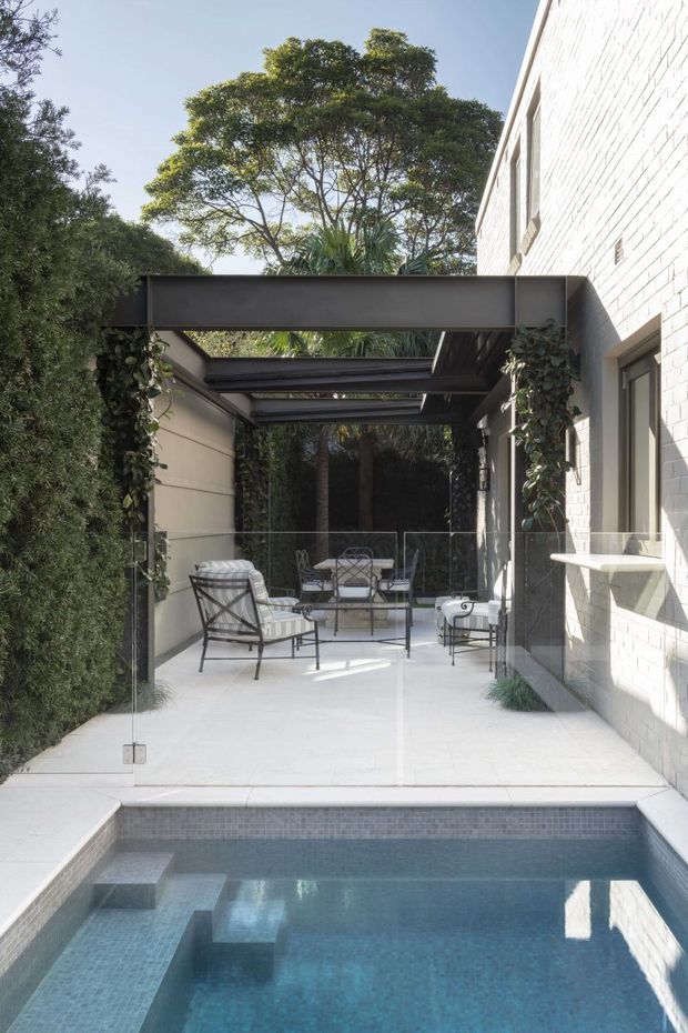 A refined outdoor courtyard terrace beside a white brick house, with a covered pergola walkway, glass balustrade, pale paving, outdoor lounge furniture, climbing greenery, and a small tiled swimming pool in the foreground. The space is bright, leafy, and sheltered between the building and dense landscaping.