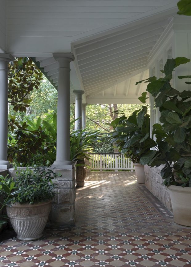 A covered veranda or porch surrounded by lush planting, featuring white-painted timber columns with classical capitals, a detailed white timber-lined pitched ceiling, white balustrades, stone column bases and low walls, and an ornate geometric encaustic-tile floor in muted red, cream, grey and brown tones.