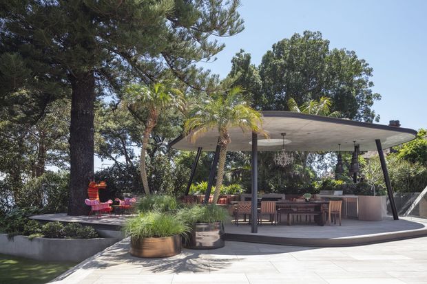 A landscaped outdoor entertaining area with a large flat-roofed pavilion, open sides, dark slender structural posts, and a built-in outdoor kitchen or bar. The pavilion includes a dining table, bar stools, pendant lighting, and warm timber decking, surrounded by mature trees, palms, raised planters, and a paved terrace.