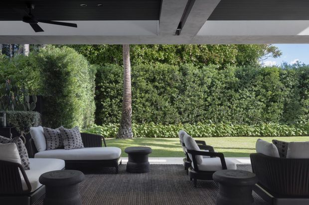 A covered outdoor lounge terrace with upholstered sofas, armchairs, low round stools and a dark ceiling fan. The space opens onto a manicured lawn bordered by dense hedges and tropical planting, creating a sheltered garden entertaining area.