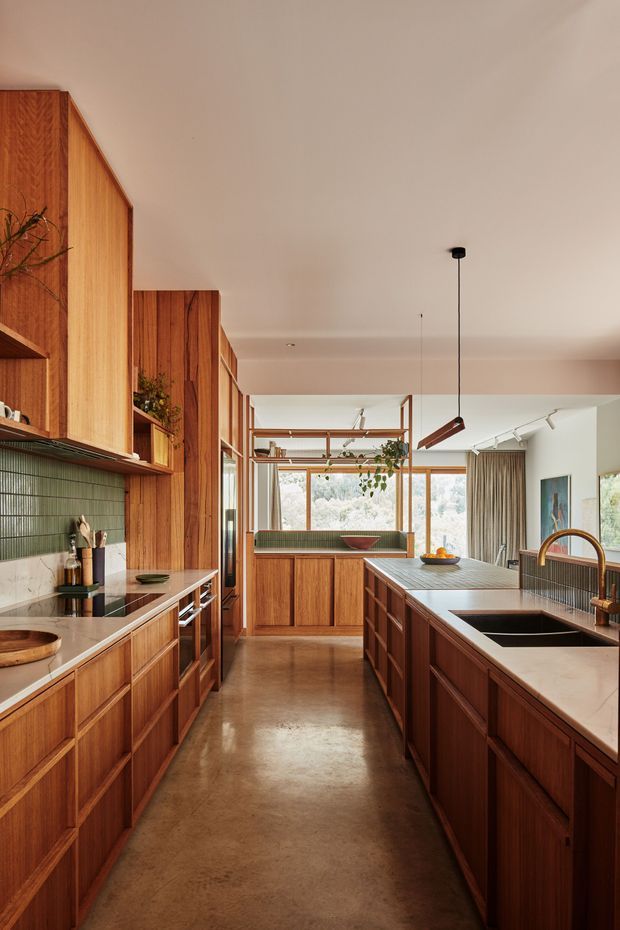 Warm, mid-century-inspired kitchen with extensive vertical timber cabinetry, a long central island, integrated cooktop, undermount sink and bronze-toned tapware. The kitchen opens toward a bright dining or living area with large windows, curtains and indoor planting. A polished concrete floor, green tiled splashback and simple pendant and track lighting create a refined, earthy interior.