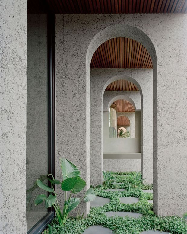 A dramatic architectural corridor with repeated rounded arch openings, rough light-grey concrete walls, dark timber slatted ceilings, and lush planted groundcover with stepping stones. Glazing along the left reflects the garden and arches, creating a layered, rhythmical perspective with a calm tropical courtyard atmosphere.