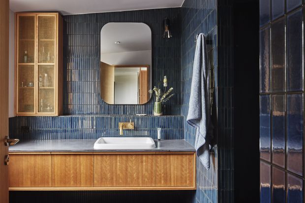 A compact, richly detailed bathroom with deep blue vertically ribbed wall tiles, a timber floating vanity with integrated basin and brass tapware, a rounded rectangular mirror, and a built-in timber and glass display cabinet. A dark framed glazed door or window is visible at right. The space has a refined mid-century-inspired character with warm timber contrasting against glossy blue tile.