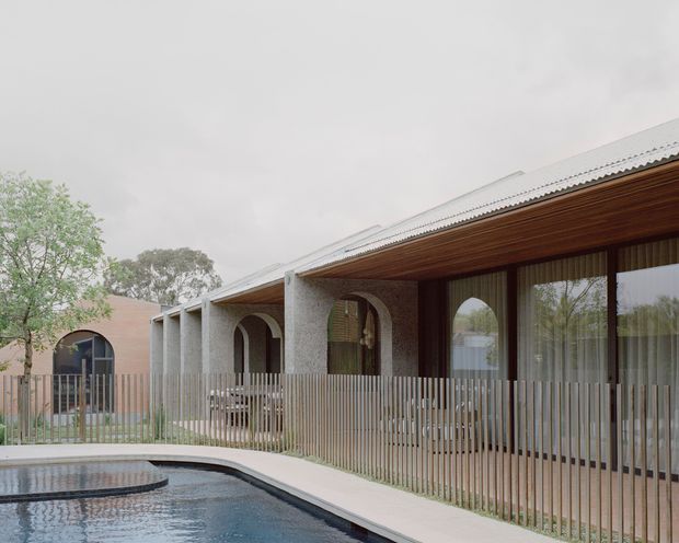 Single-storey contemporary residential building viewed from the pool area, with a long low-pitched corrugated roof, deep timber-lined eaves, textured light masonry walls, arched openings, extensive glazed doors, and a vertical metal picket fence enclosing the foreground terrace. A curved swimming pool and paved surround occupy the front of the image, with mature trees visible beyond.