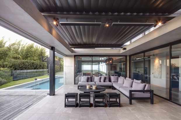Covered outdoor living area beside a swimming pool, featuring a large sectional sofa and coffee tables, dark metal roof decking, exposed concrete beams, slim dark-framed glazing, and a timber pool deck. The space opens toward landscaped grounds and water views, creating a refined indoor-outdoor entertaining area.