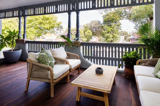 A covered, elevated timber veranda furnished as an outdoor lounge, with rattan armchairs and sofas, white cushions, a slatted timber coffee table, potted tropical plants, dark timber decking, and decorative dark-painted balustrades and posts. The open sides overlook a leafy suburban setting.