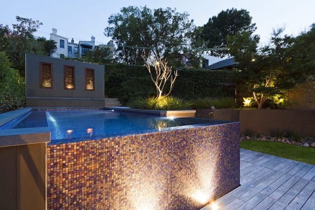 A landscaped residential pool courtyard photographed at dusk, featuring a raised rectangular swimming pool with a mosaic-tiled exterior and illuminated water feature, grey tiled paving, sculptural planting, hedges, and a dark rendered feature wall with narrow lit windows. Modern residential buildings are partially visible behind the garden.