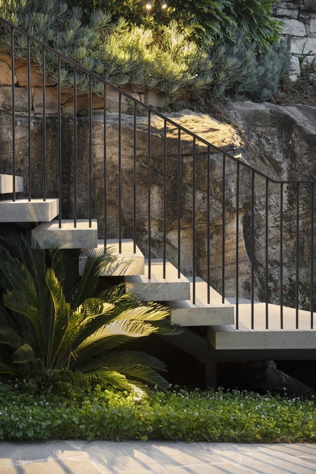 Exterior view of a modern concrete stairway integrated into a rocky landscaped garden. The stair has floating concrete treads and slim black metal vertical balustrades, with dense planting and exposed natural stone forming the backdrop.