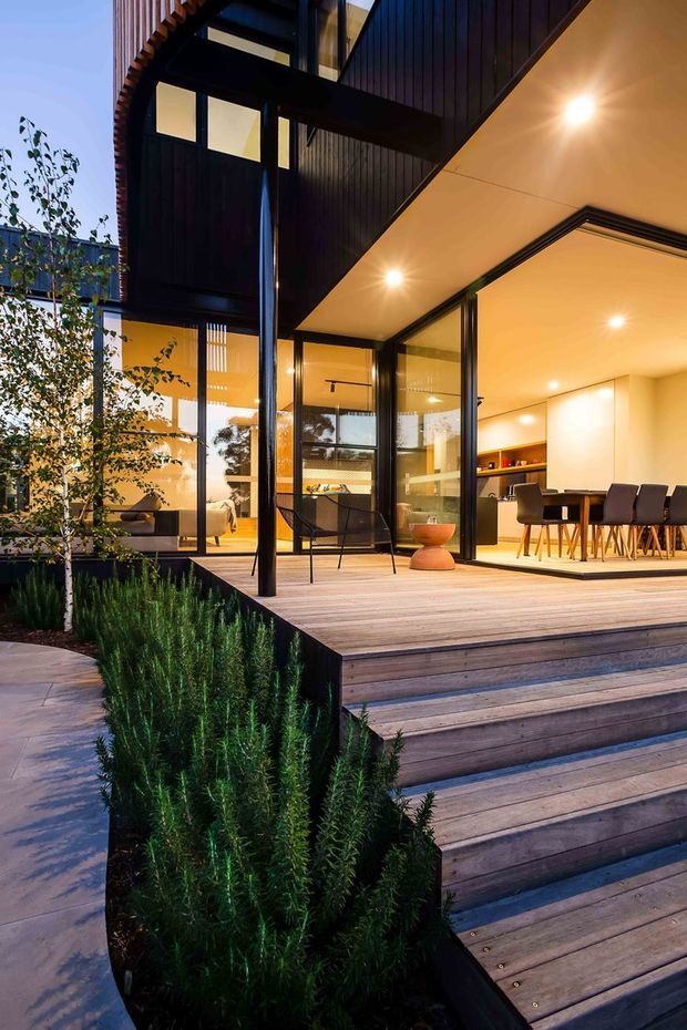 Contemporary multi-level home viewed from a landscaped timber deck and stairway at dusk. The design features extensive floor-to-ceiling glazing, dark vertical timber cladding, black-framed doors and windows, deep roof overhangs, and warm recessed interior lighting. The deck opens into a bright dining and kitchen area, with a living space visible beyond the glazing. Linear planting borders the timber stairs and terrace.