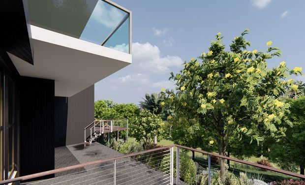 Exterior view of a contemporary multi-level house showing a projecting upper balcony with clear glass balustrading, a lower deck with horizontal metal cable railing, dark vertical cladding, and lush landscaped vegetation beyond.
