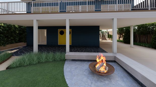 A landscaped courtyard beneath a raised contemporary home, with white structural columns, dark horizontal cladding, an upper-level balcony with metal balustrade, and a central outdoor fire bowl set on irregular stone paving. Artificial lawn, ornamental grasses, hedges, and pale built-in bench seating frame the outdoor entertaining area.