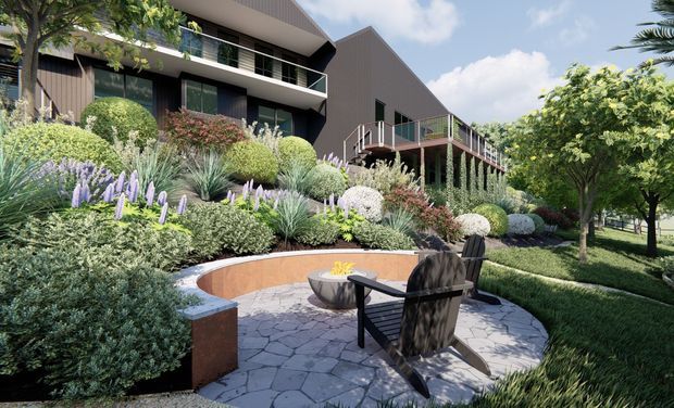 A landscaped outdoor courtyard and seating area in front of a contemporary multi-level residential building. The garden features layered ornamental planting, curved raised garden beds, stone-paved paving, timber outdoor chairs, and a small fire bowl. The building has dark vertical cladding, broad glazing, balconies, and a prominent pitched/gabled roof form.