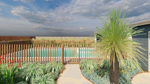 A landscaped outdoor pool area viewed through a vertical timber slat fence, with turquoise pool water, pale paving, dense ornamental grasses and a sculptural yucca-like plant. A dark-clad building edge is visible at right beneath a dramatic partly cloudy sky.