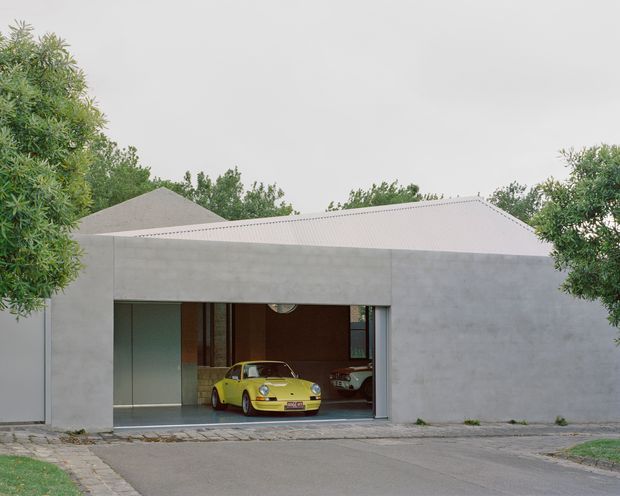 A low, minimalist residential garage or carport facade with smooth grey concrete walls and a shallow pitched metal roof. A wide open garage reveals a bright yellow classic sports car and another vehicle inside, with mature trees surrounding the property and a paved driveway in front.