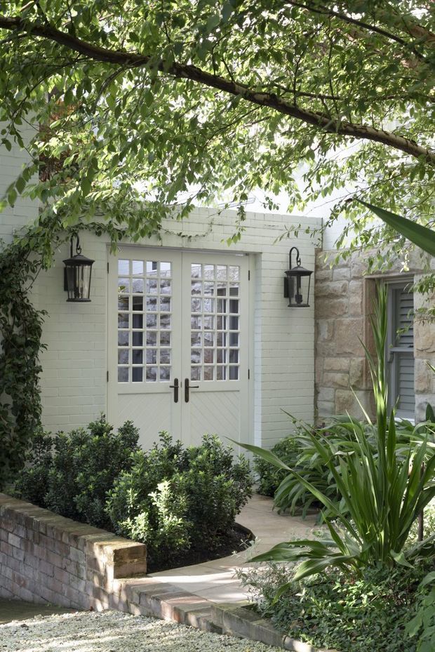 A leafy landscaped entry courtyard leading to pale painted French-style double doors with multi-pane glazing. The entrance is framed by black wall lanterns, clipped shrubs, tropical planting, a low stone retaining wall, and a natural stone masonry wall, creating a sheltered traditional garden approach.