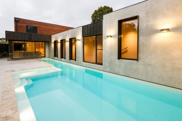 Modern flat-roofed residential house viewed from its swimming pool. The composition features a long pale rendered/concrete facade with tall dark-framed windows, warm interior lighting, black metal roof and wall elements, and a contrasting timber-clad upper volume. The turquoise pool dominates the foreground and wraps alongside the building, creating a clean, contemporary indoor-outdoor setting.