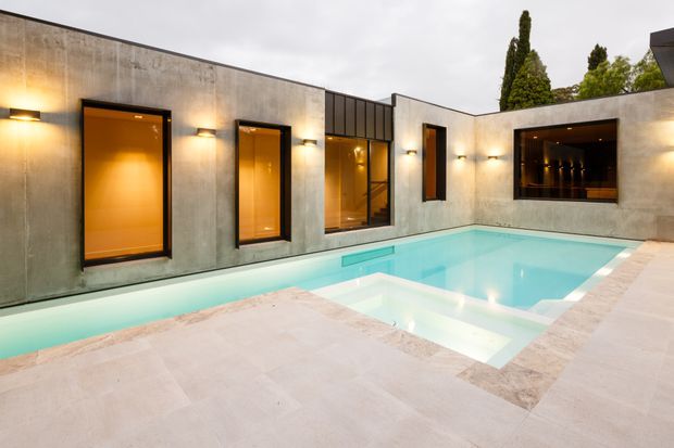 Exterior view of a minimalist modern courtyard centered on a rectangular swimming pool. The low flat-roofed building has smooth concrete walls, dark-framed windows and doors with warm interior lighting, and pale stone or concrete pool paving. A recessed outdoor bar or service opening is visible on the right wall.
