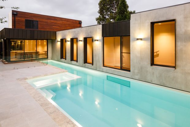 Exterior view of a contemporary residential courtyard with a long turquoise swimming pool adjoining a low, flat-roofed building. The facade combines smooth pale concrete or rendered walls, dark metal cladding, large black-framed windows, and a warm timber-clad upper volume. Interior lighting glows through the glazing at dusk, creating a refined indoor-outdoor setting.